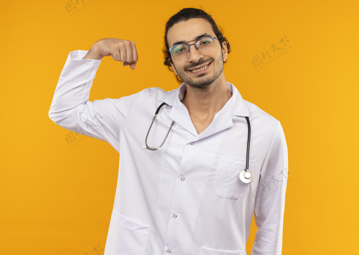 医学微笑的年轻医生戴着医用眼镜穿着医用长袍用听诊器在黄色上做着强烈的手势长袍强壮空间