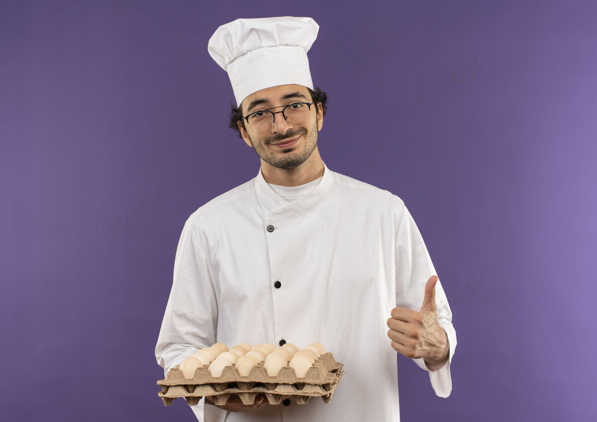 男性高兴的年轻男厨师穿着厨师制服 戴着眼镜 手里拿着一批鸡蛋 大拇指朝上放在紫色上拇指烹饪紫色