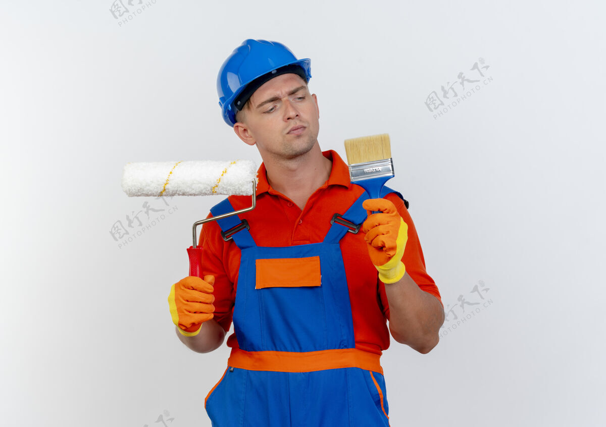 制服想着年轻的男建筑工人穿着制服 戴着安全帽 手里拿着油漆辊 看着油漆刷上的白色思考头盔安全