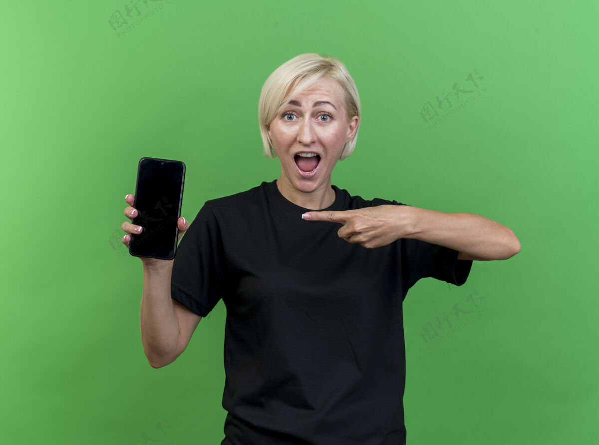 感觉印象深刻的中年金发女人看着前面展示的手机指着它隔离在绿色的墙上手机表演衣服