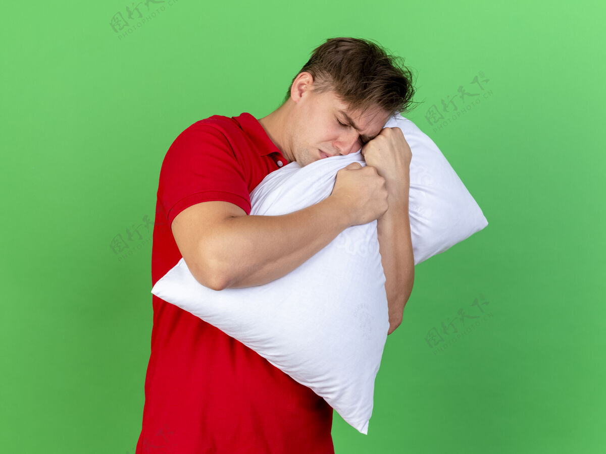 男人皱眉的年轻英俊的金发病男子抱着枕头闭着眼睛孤立在绿色的墙上脸年轻姿势