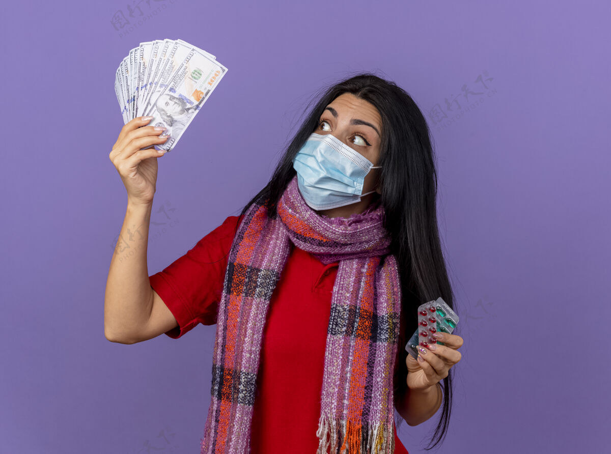 人印象深刻的年轻生病的女人戴着面具和围巾拿着钱和一包胶囊看着隔离在紫色墙上的钱脸抱衣服