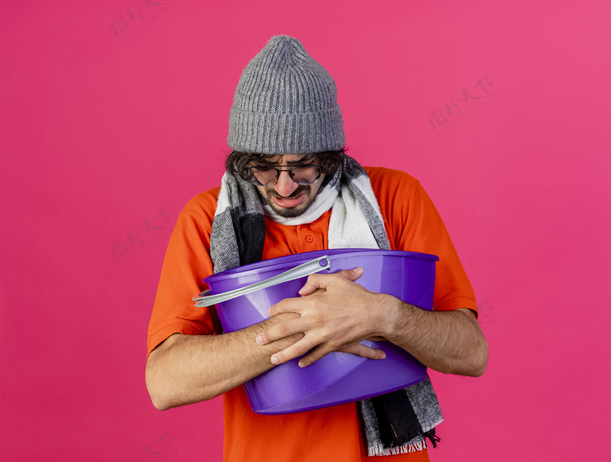帽子愁眉不展的年轻病夫戴着眼镜 戴着冬天的帽子 戴着围巾 手里拿着塑料桶 恶心地看着隔离在粉红色墙上的墙人倒眼镜