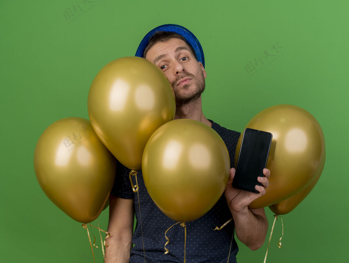 人戴着蓝色派对帽的自信帅哥手持氦气球站在绿色的墙上 把电话隔离开氦持有人