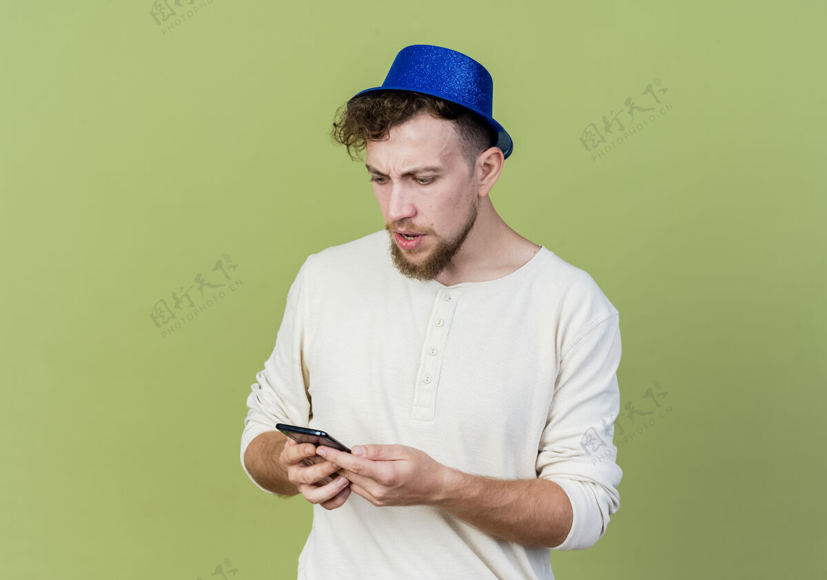 抱着愤怒的年轻帅哥戴着派对帽拿着手机看着孤零零的橄榄绿墙上站着聚会衣服