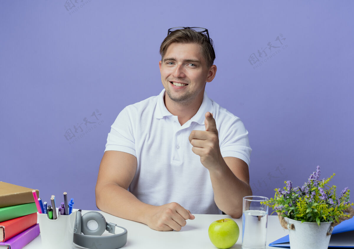 帅气微笑的年轻英俊的男学生坐在书桌旁 手里拿着学习用具 头戴眼镜 在蓝色的舞台上向你展示孤立的姿势学校头书桌
