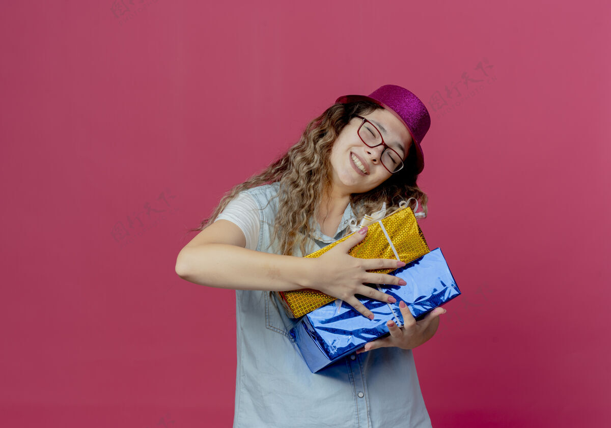 年轻闭上眼睛微笑的年轻女孩戴着眼镜 戴着粉红色的帽子 手里拿着孤立的粉红色礼盒帽子礼物佩戴