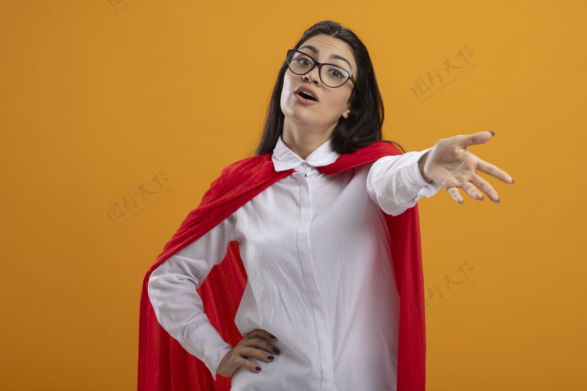 伸展年轻的女超人戴着眼镜望着前面 伸出一只手朝前 把另一只手放在腰上 隔离在橙色的墙上保持年轻表情