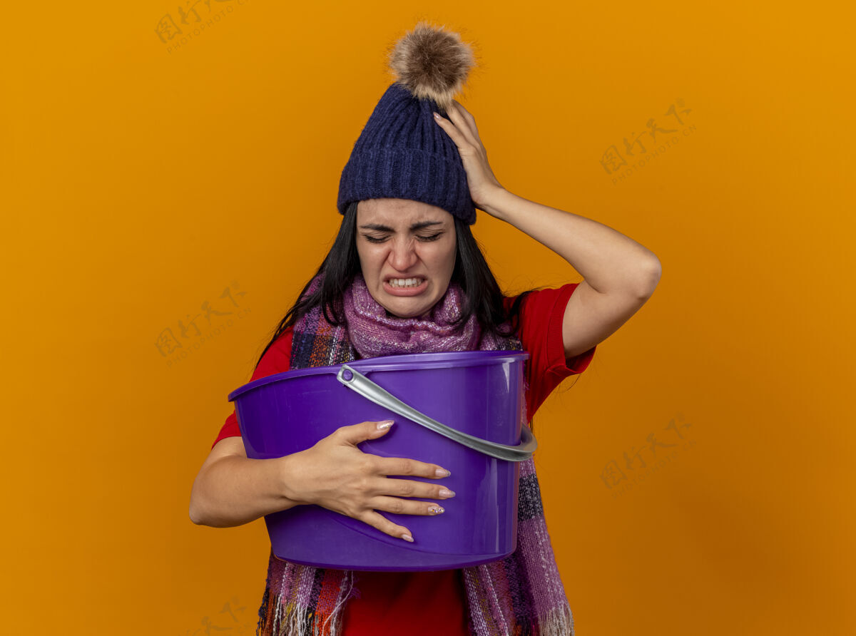 保持恼怒和疼痛的年轻患病妇女戴着冬天的帽子和围巾拿着塑料桶恶心保持手放在头上闭着眼睛隔离在橙色的墙上市民冬天疾病