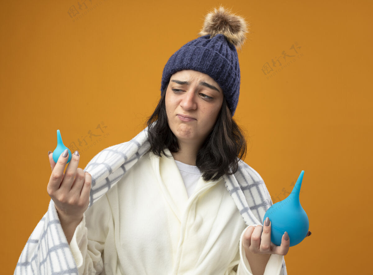 表情可疑的年轻生病的女人穿着长袍 戴着格子布裹着的冬帽 拿着灌肠剂 看着隔离在橙色墙上的小家伙长袍站疾病