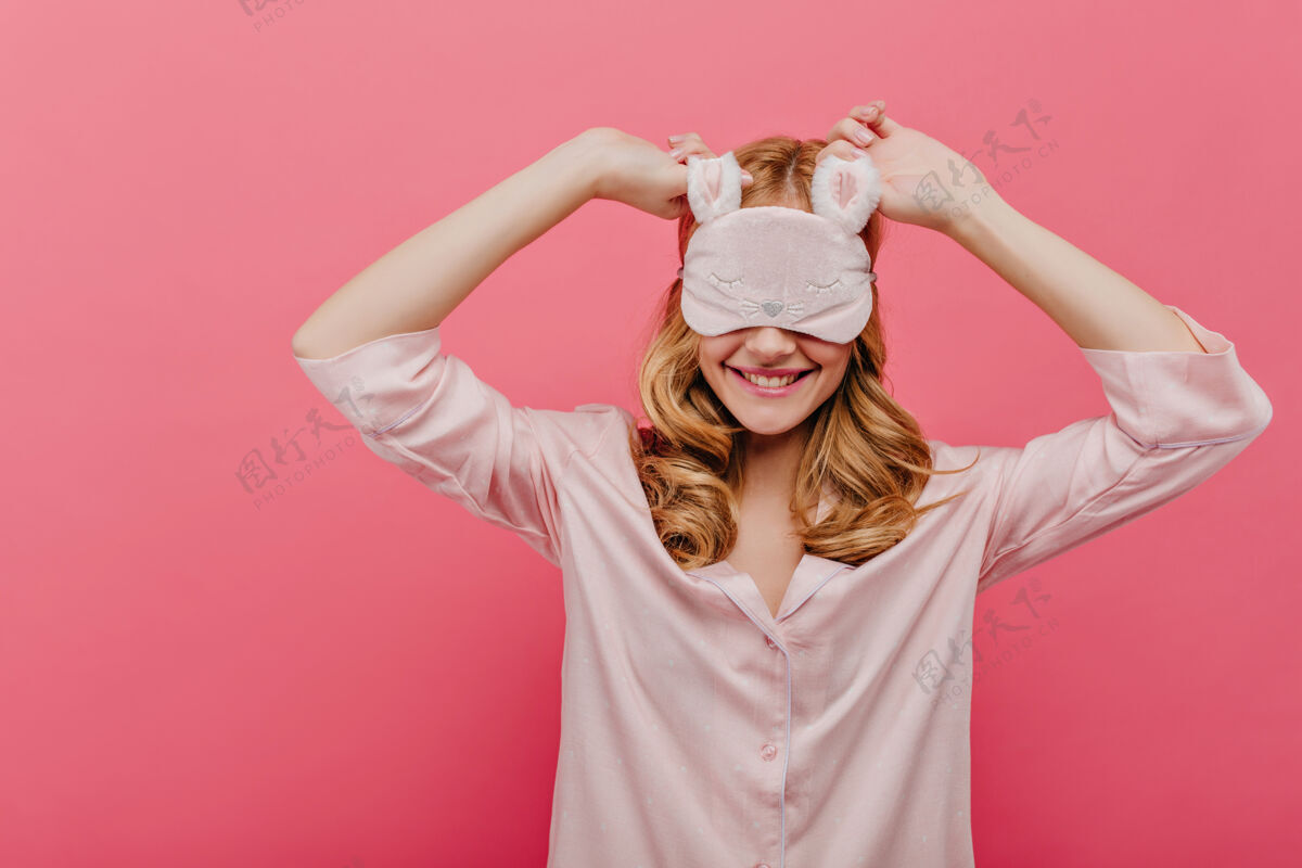装备迷人的卷发女孩戴着眼罩 在周末的早晨欢笑幸福的白色女模特穿着丝绸睡衣在粉色的墙上嬉戏成人服装眼罩