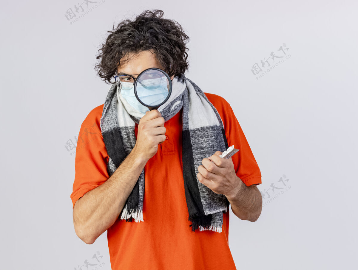 衣服一个戴着眼镜 围巾和口罩的年轻人拿着一包包药片 透过隔离在白墙上的放大镜看着他们疾病人医学