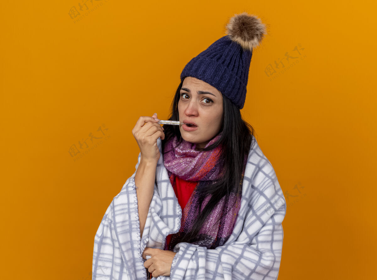 穿着印象深刻的年轻生病的女人戴着冬天的帽子和围巾裹在格子布把温度计放在嘴里看着前面孤立的橙色墙壁人站立表情