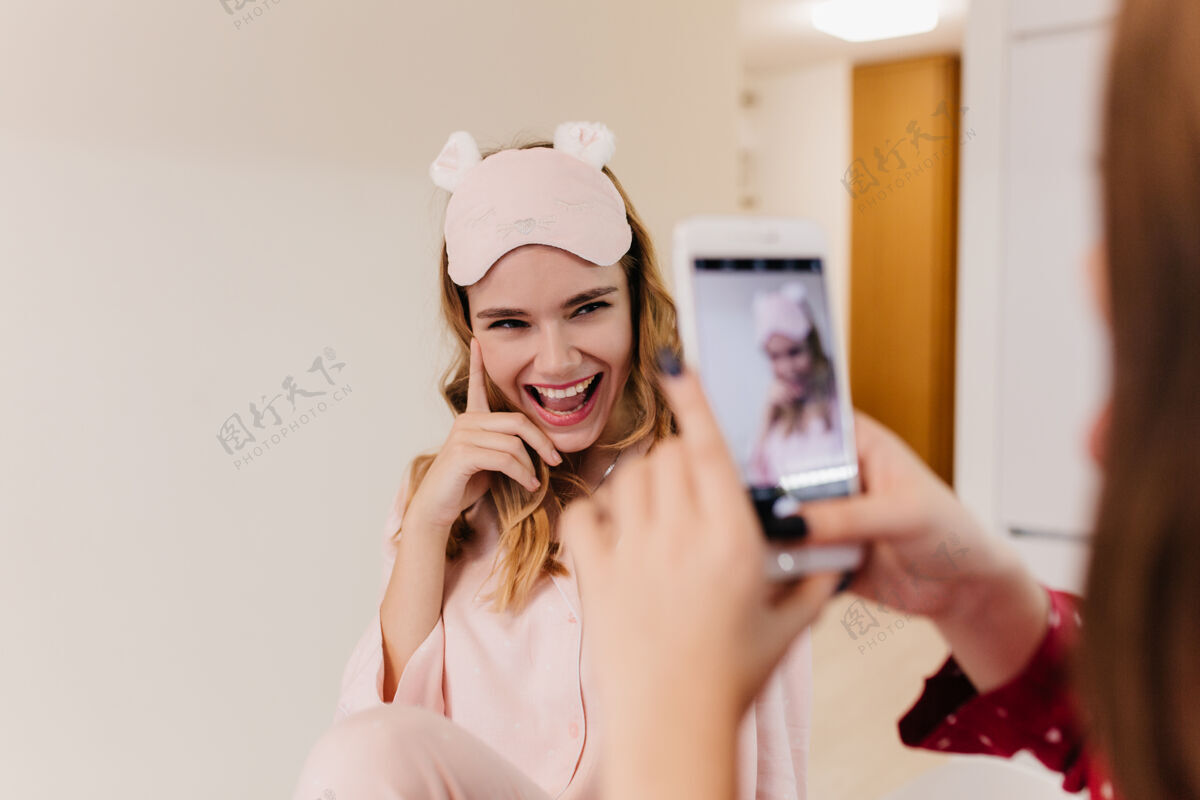手机一个白人女孩在姐姐面前摆出俏皮的微笑一个黑发女人拿着手机给戴着粉红色眼罩的朋友拍照快乐微笑享受