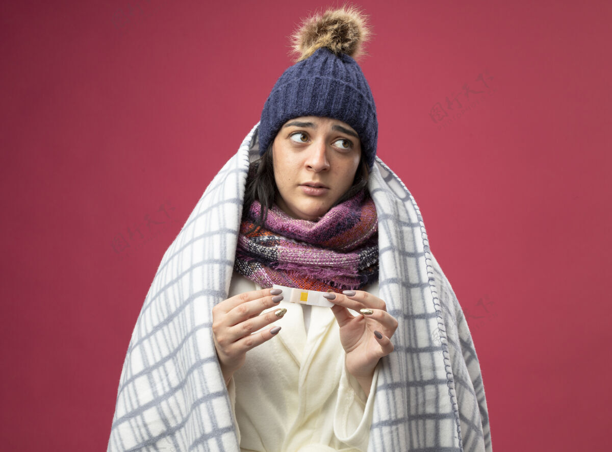 感情给人印象深刻的年轻生病的女人穿着长袍 戴着冬天的帽子 围着方格布裹着的围巾 手里拿着医用石膏 看着隔离在粉红色墙上的一面衣服人人