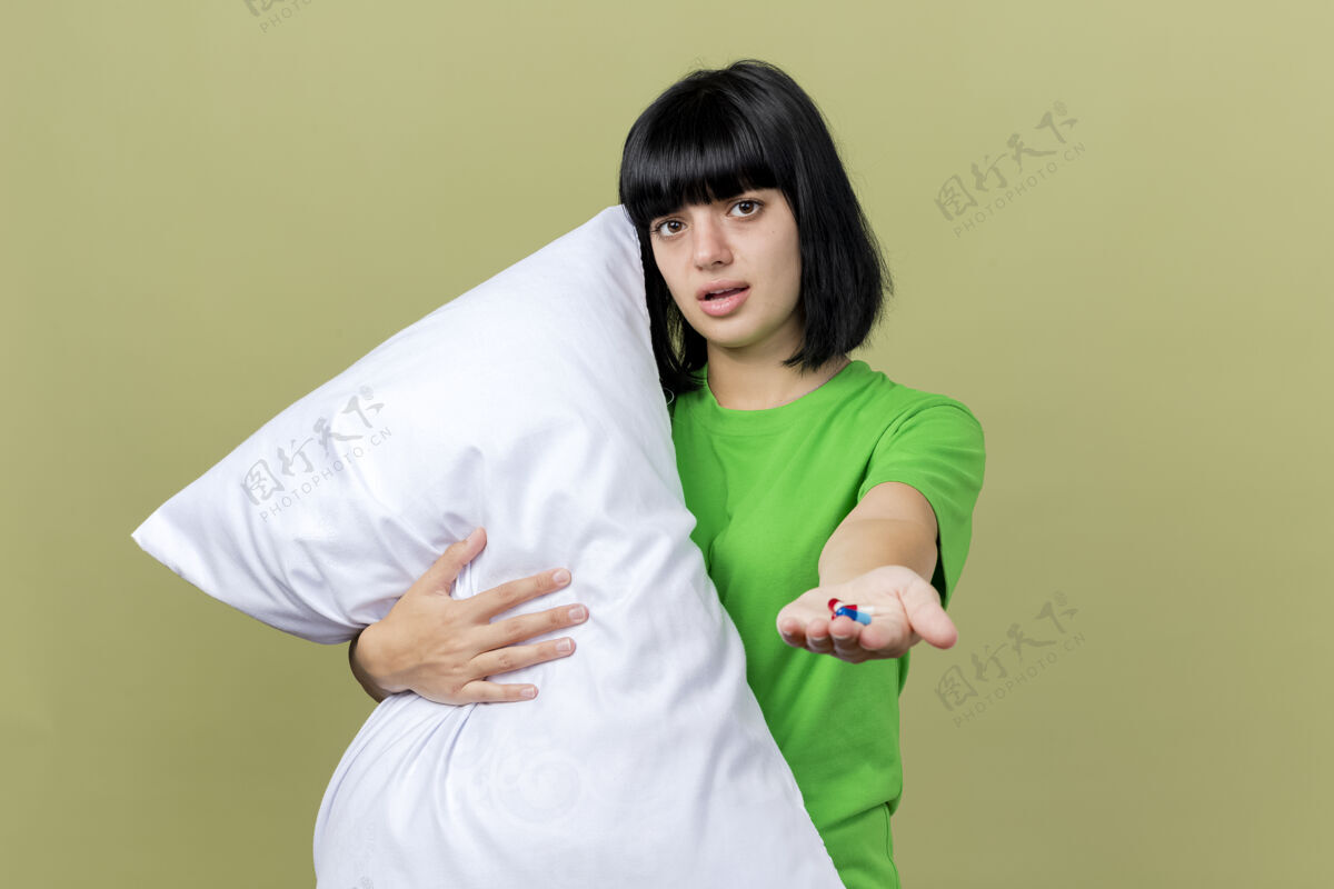 抱着虚弱的年轻病妇抱着枕头看着前面伸出的医药胶囊 朝着前面隔离在橄榄绿的墙上虚弱姿势衣服