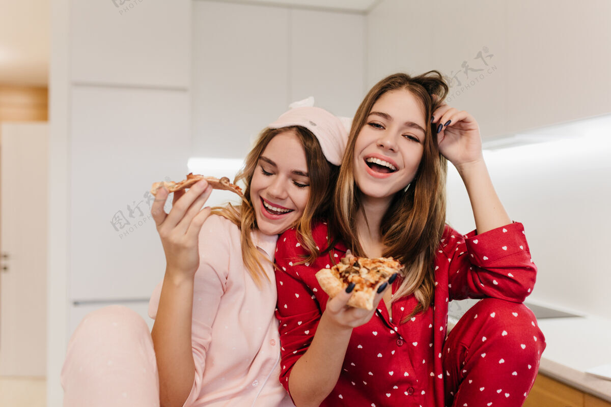 娱乐穿着可爱睡衣的欧洲女人和戴着眼罩的妹妹一起欢笑美丽的白人女士愉快地吃着奶酪披萨微笑室内快乐