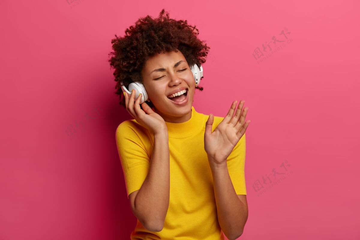 罗西喜出望外的无忧无虑的女人的肖像听音乐 唱歌戴着耳机 闭上眼睛 忘记所有的问题 穿着黄色的衣服 隔离在粉红色的墙上生活方式的概念女性微笑旋律