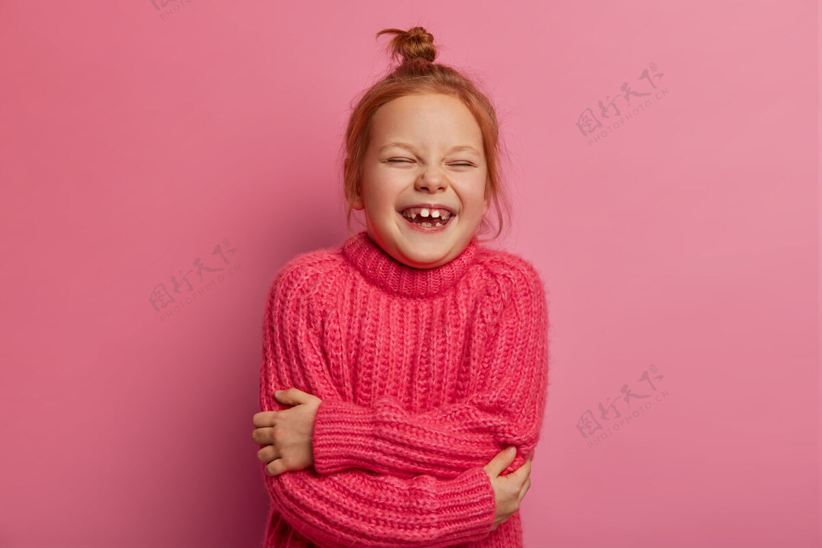 欢笑喜出望外的姜汁小女孩搂着自己 有着积极的表情 穿着温暖的针织毛衣 喜欢拍照 表达真挚美好的情感 孤立在粉色的墙上孩子们 娱乐快乐休闲温暖