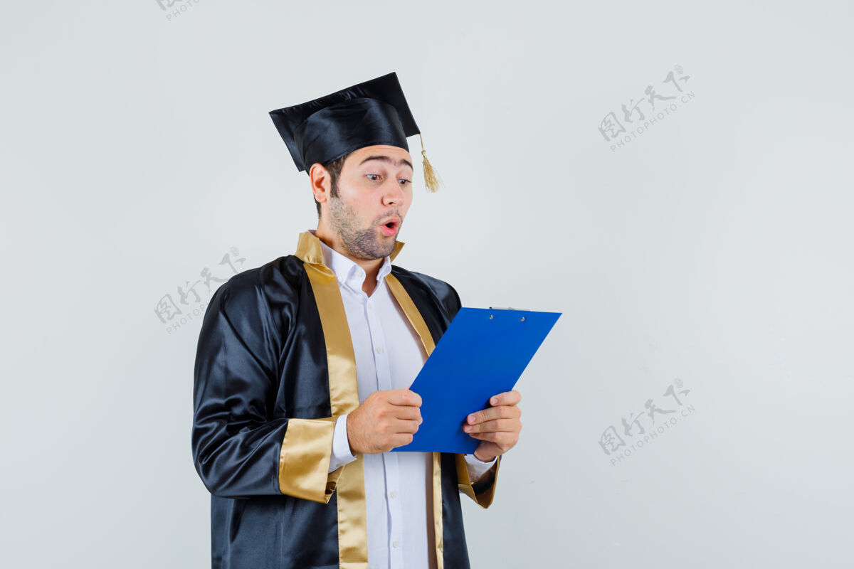 男人年轻人穿着毕业制服 看着剪贴板上的笔记 惊讶地看着前方的景色剪贴板前面欢呼