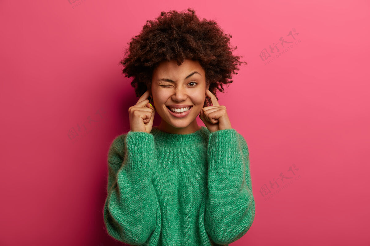 肖像漂亮女人的画像塞住耳朵眨眼睛 笑容灿烂 无视不愉快的嘈杂声 穿着绿色毛衣 对着粉色的墙壁摆姿势 表情愉快关掉音量乐观模特请