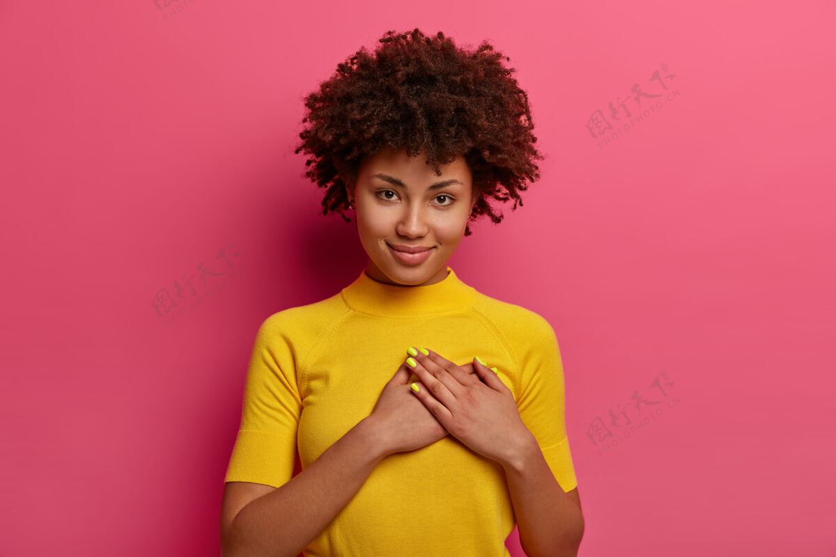 女人可爱的卷发女人把手掌放在心上 感激礼物 穿着黄色t恤 在玫瑰色的墙上摆姿势 说你永远在我心里 温柔地微笑自信感谢种族