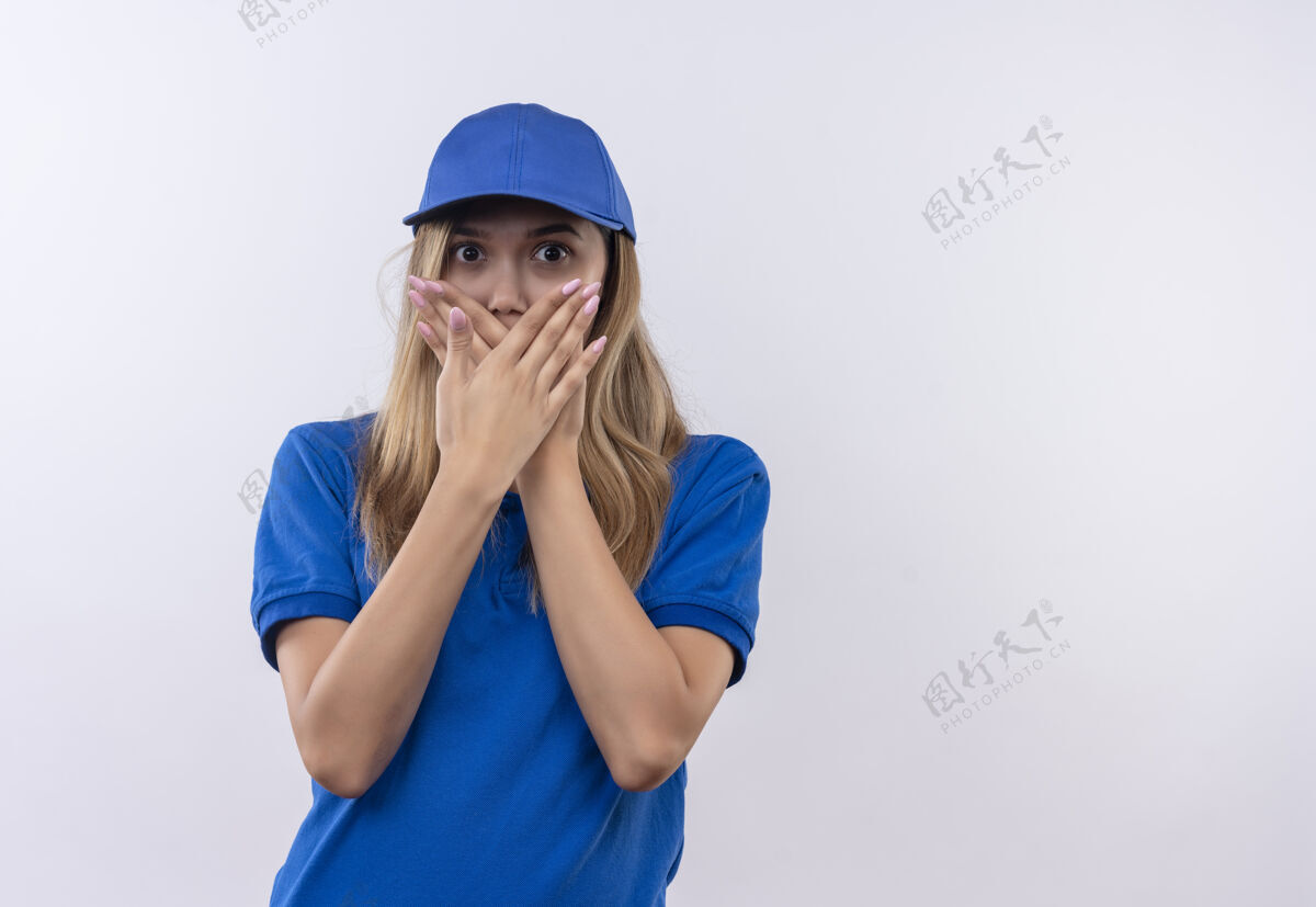 表情害怕的年轻送货女孩穿着蓝色制服 戴着帽子 嘴巴被隔离在白色的手上手手势害怕
