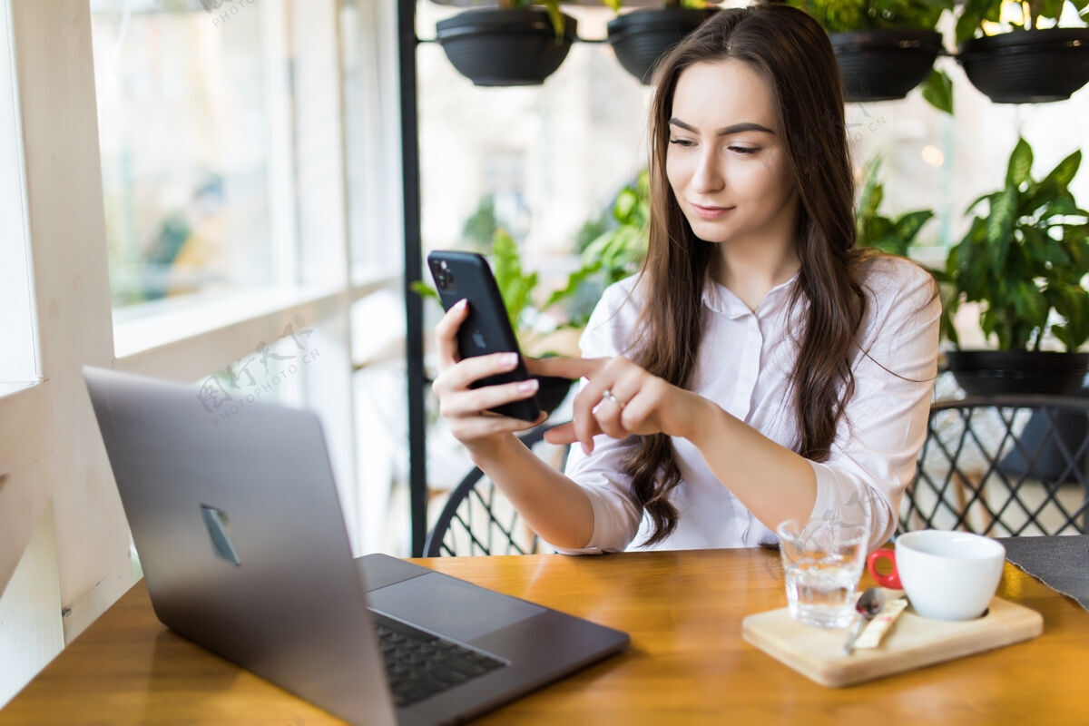 休闲年轻漂亮的金发女郎拿着手机 坐在咖啡厅里面拿着便携上网本 迷人的梦幻女郎在咖啡厅休息时用手机和笔记本电脑网络电话应用程序