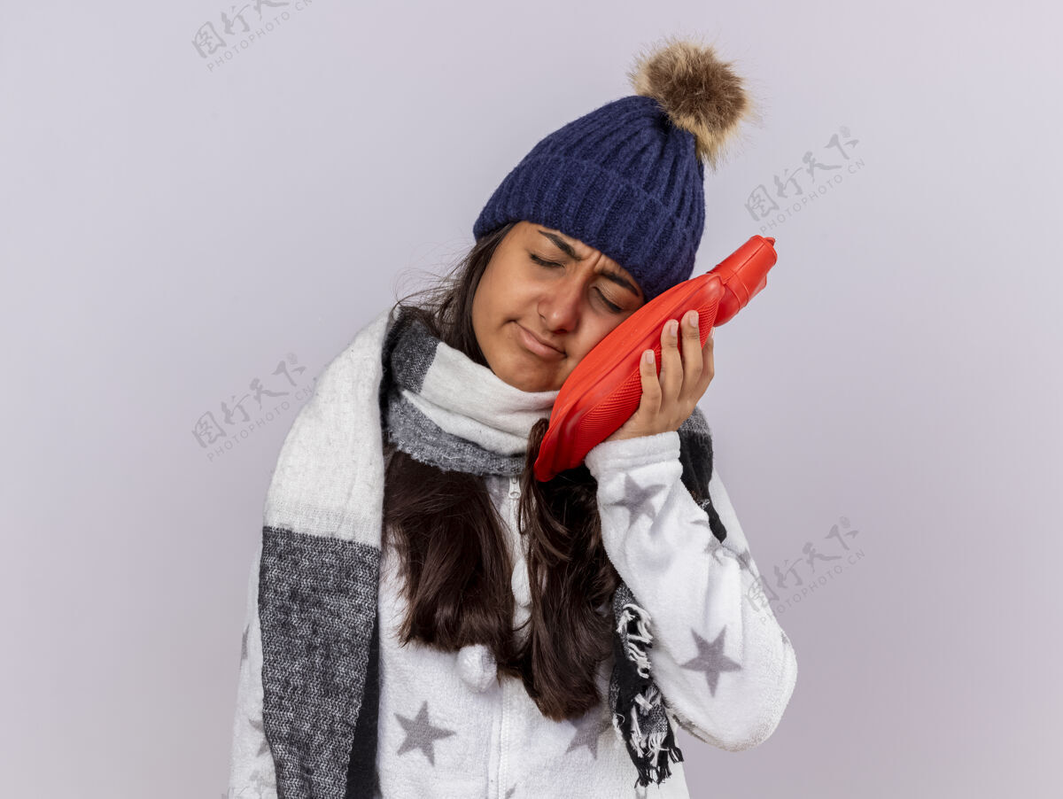 悲伤悲伤的年轻病女孩闭着眼睛戴着冬天的帽子戴着围巾把热水袋放在脸颊上隔离在白色背景上脸颊包女孩