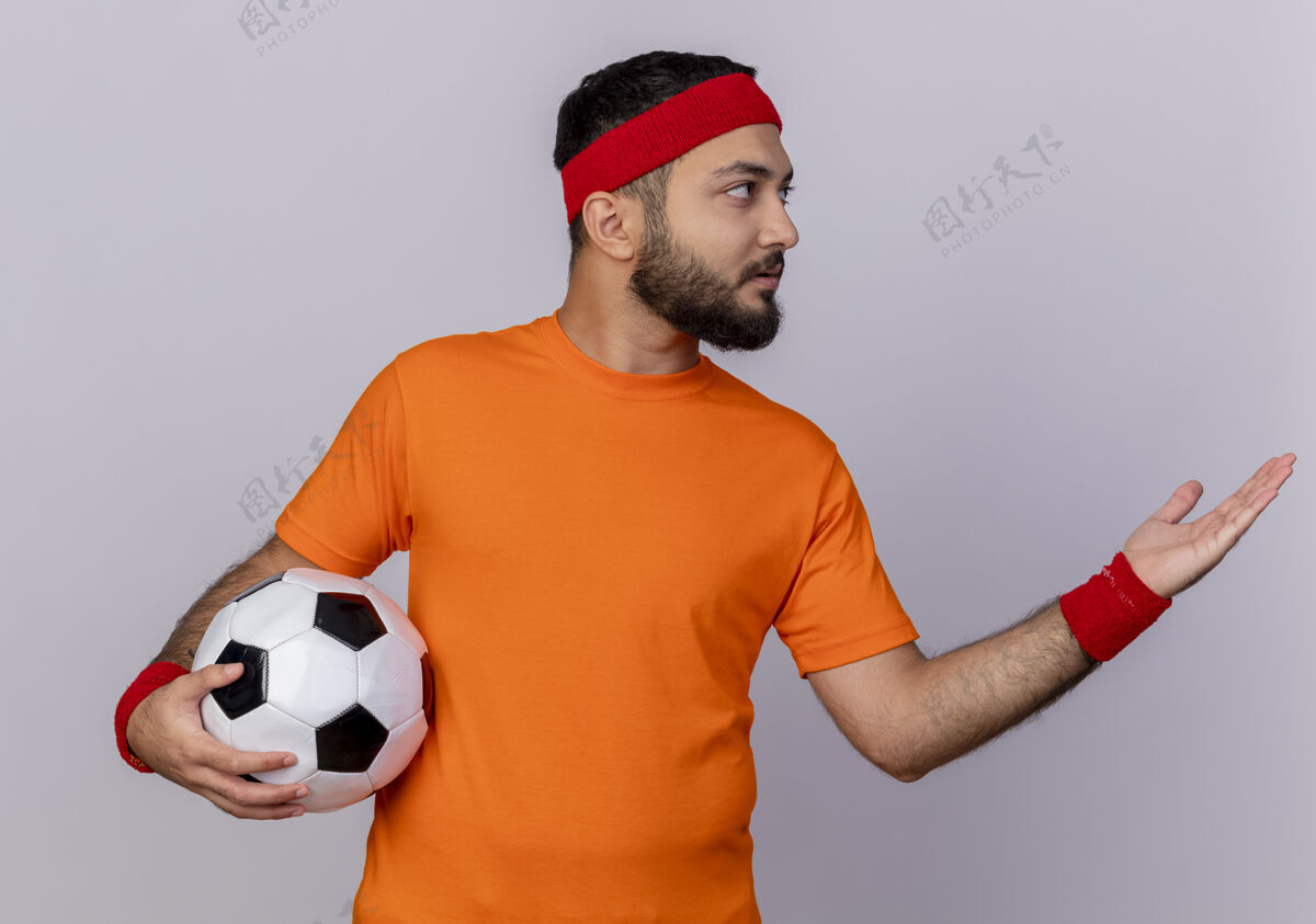 手年轻的运动型男子一边看着戴着头带和手环拿着球 一边用手指着隔离在白色背景上的一边壁板头带球
