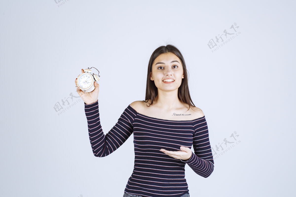 年轻人穿着条纹衬衫的女孩手里拿着闹钟 把它当作一种新产品来推销成人女售货员姿势