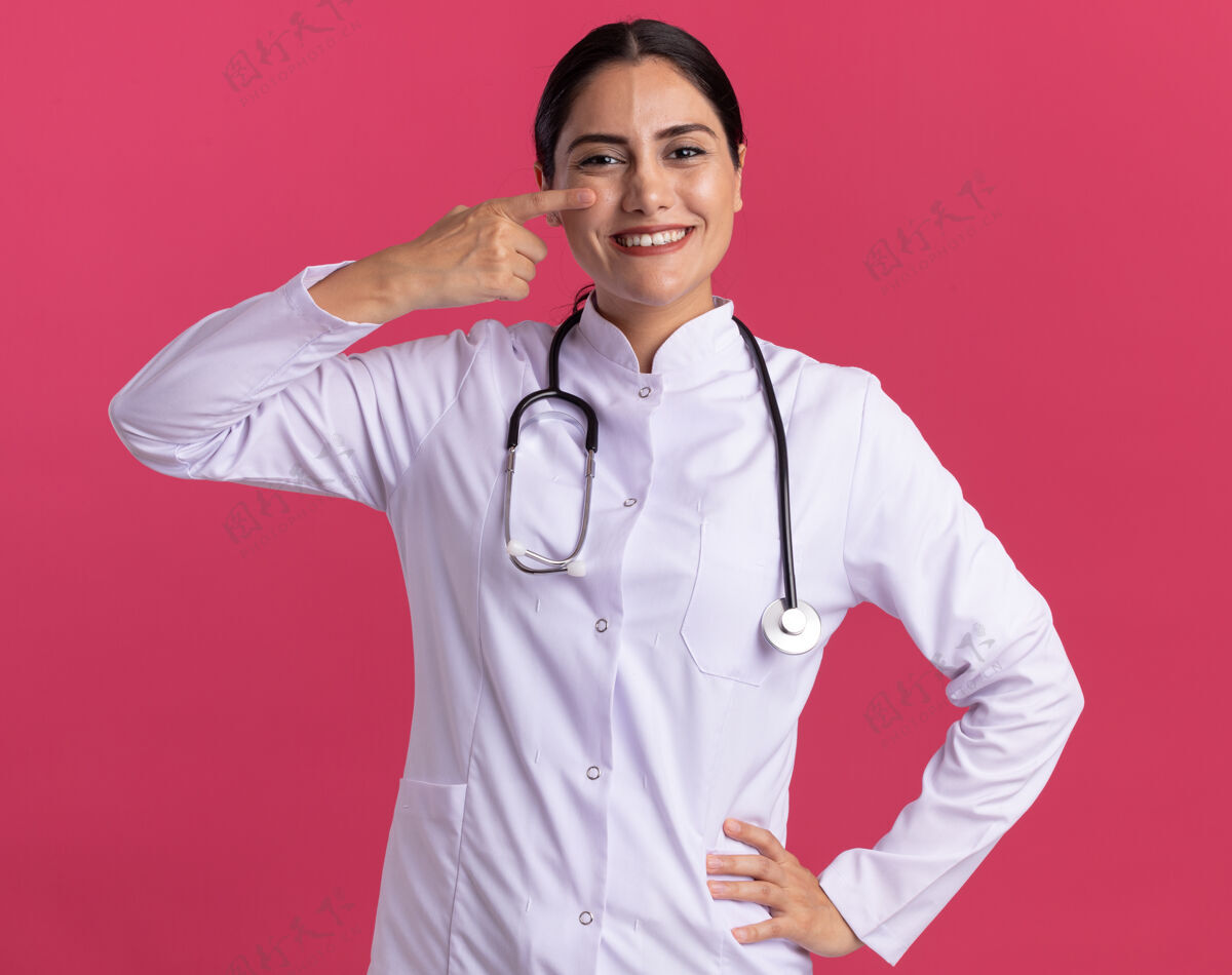 听诊器年轻的女医生穿着医用外套 拿着听诊器 看着前面 用食指指着眼睛 站在粉红色的墙上 脸上露出幸福的笑容快乐医生目录
