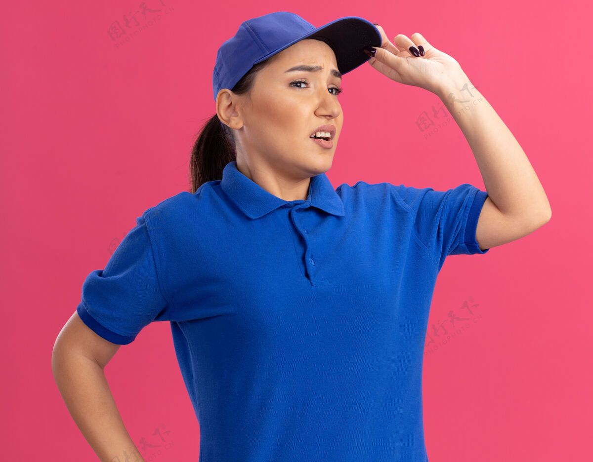 年轻身穿蓝色制服 戴着帽子的年轻送货员站在粉红色的墙上 茫然地向一旁望去制服帽子站立