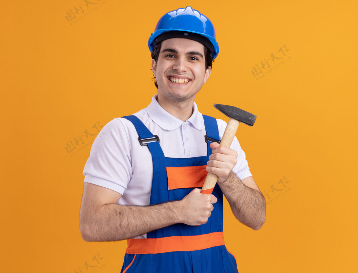 站着快乐的年轻建筑工人身穿建筑制服 戴着安全帽 手持铁锤 站在橘色的墙上 面带微笑地看着前方建筑男人安全