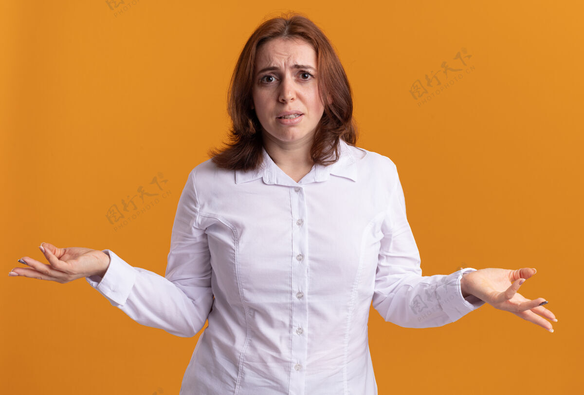 年轻身穿白衬衫的年轻女子困惑地看着前面 两臂伸向两边站在橙色的墙上女人衬衫站立