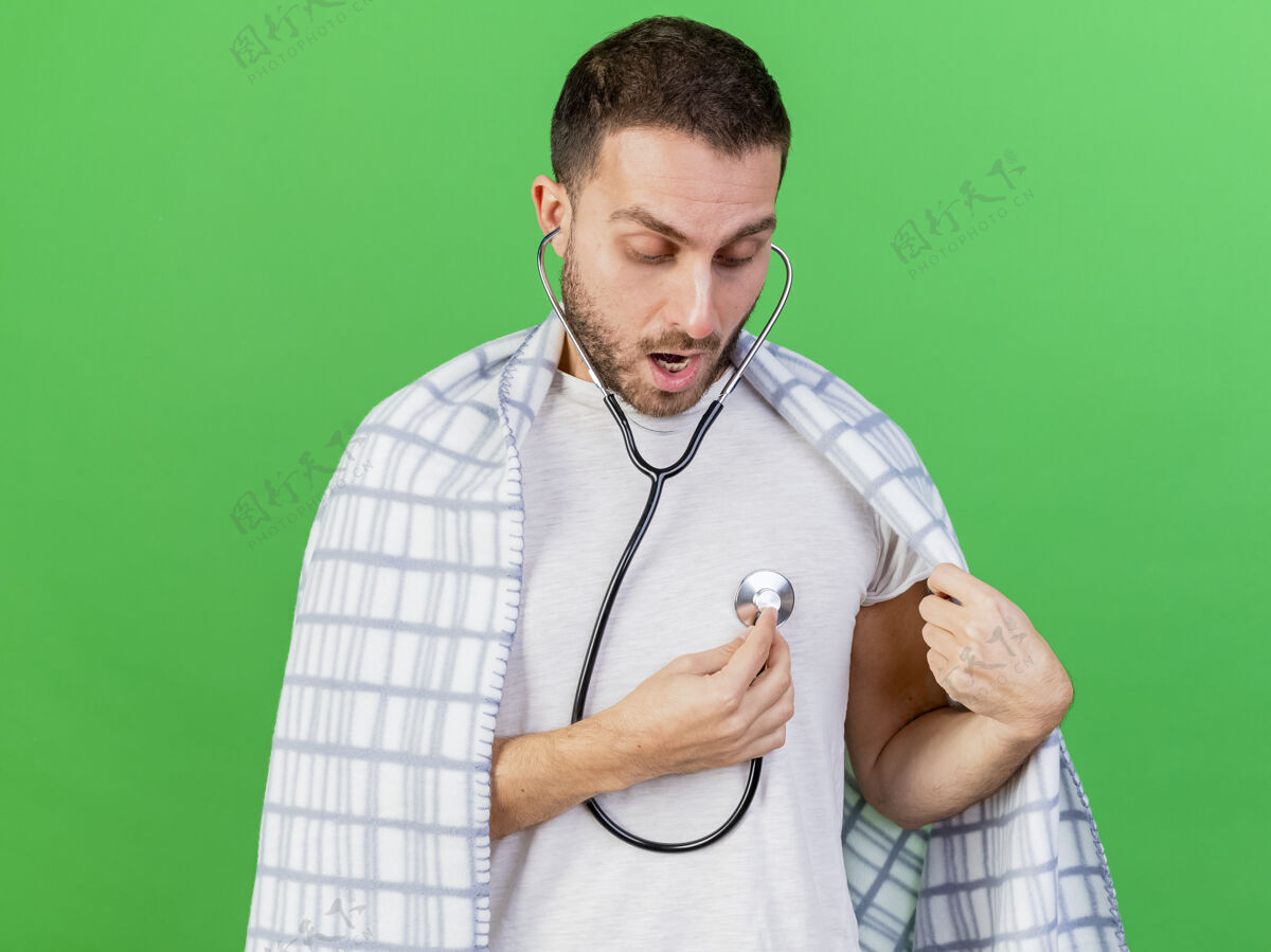 年轻一个穿着格子花呢衣服的年轻人惊讶地站在绿色的背景上 用听诊器听自己的心跳包自己的格子