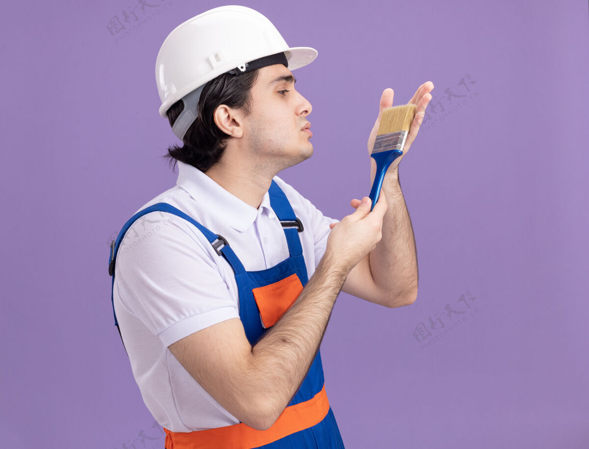 爱年轻的建筑工人穿着建筑制服 戴着安全帽 拿着油漆刷 站在紫色的墙上 带着爱看着它男人站着安全