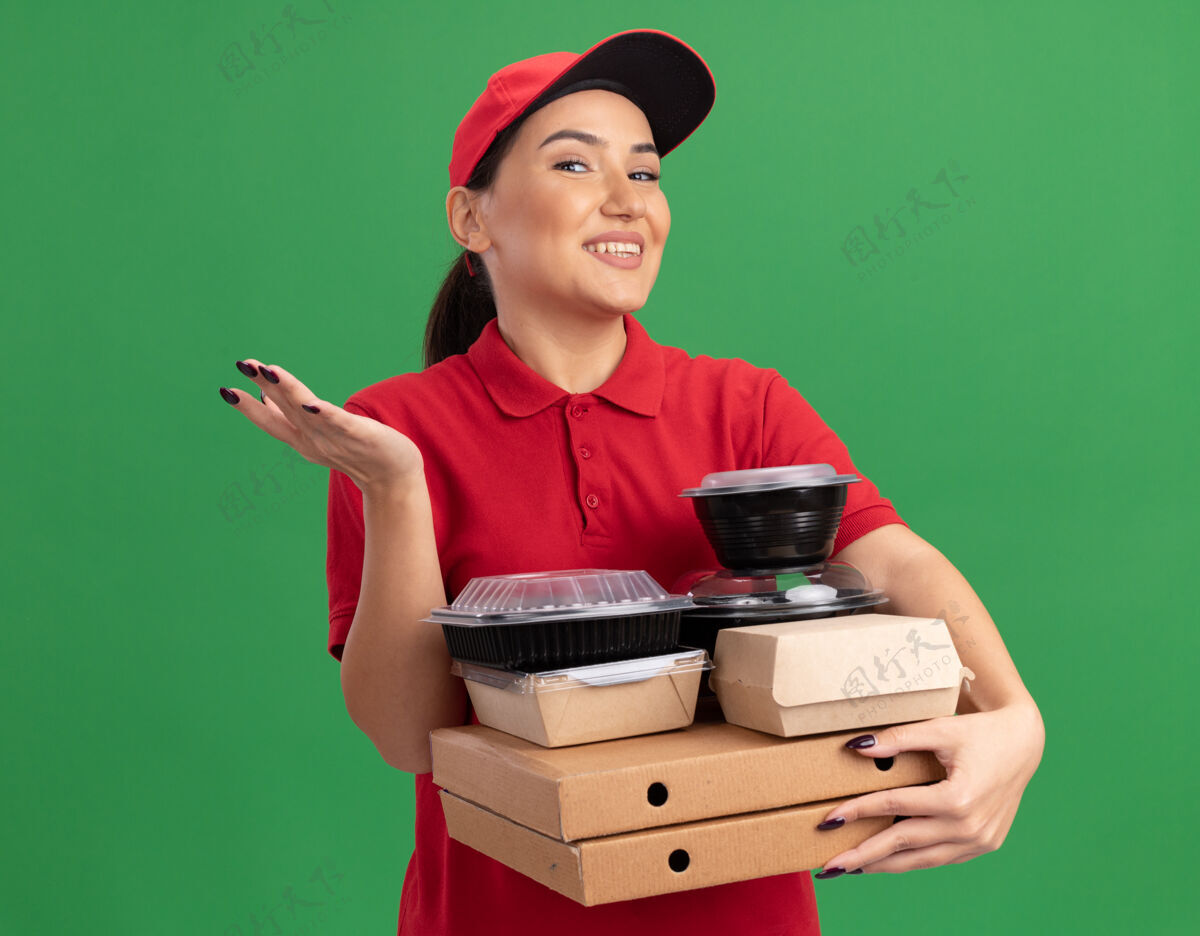 披萨身穿红色制服 头戴鸭舌帽的年轻女送货员举着比萨饼盒和食品包 面带微笑地看着前面 手臂举过绿色的墙壁微笑制服举起