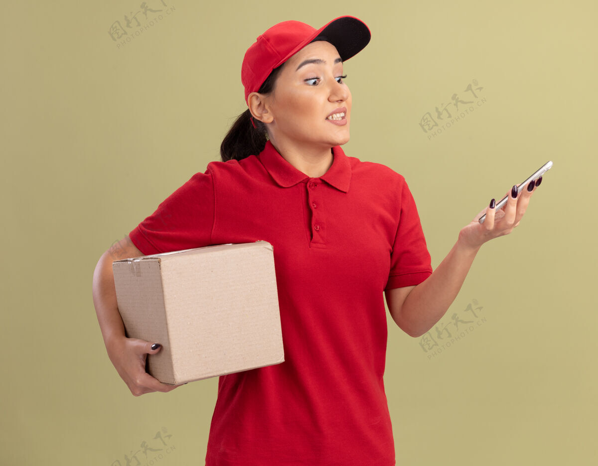 帽子年轻的送货员身穿红色制服 戴着帽子 手里拿着纸板箱 站在绿色的墙上 看着自己的智能手机 感到困惑和不快纸板移动困惑
