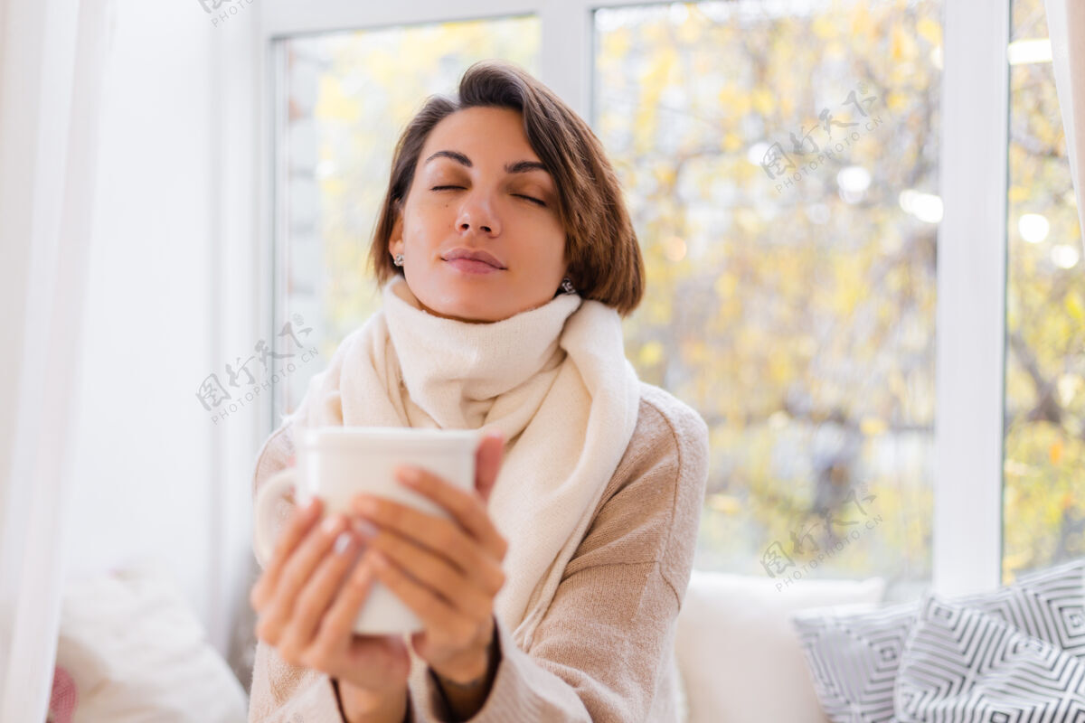 热坐在窗台上喝着热茶喝着咖啡 穿着毛衣和白围巾的女人的温馨肖像季节室内早餐