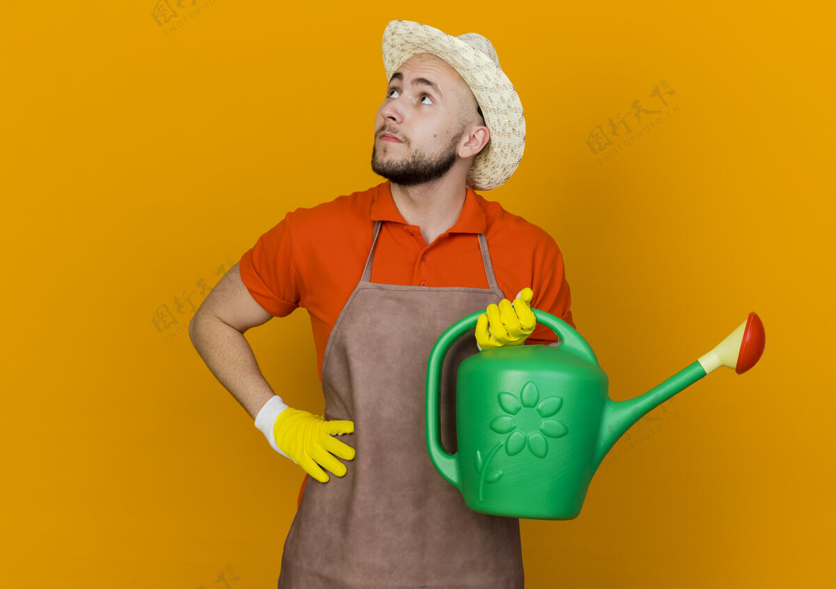 复制年轻的男园丁戴着园艺帽 把手放在腰上 拿着浇水罐看着旁边空间穿橙色