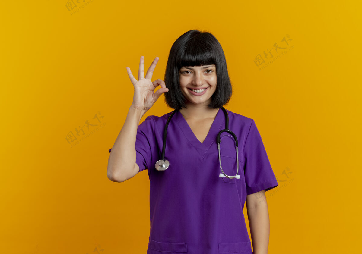 标志微笑的年轻黑发女医生穿着制服用听诊器手势ok手势黑发好女性