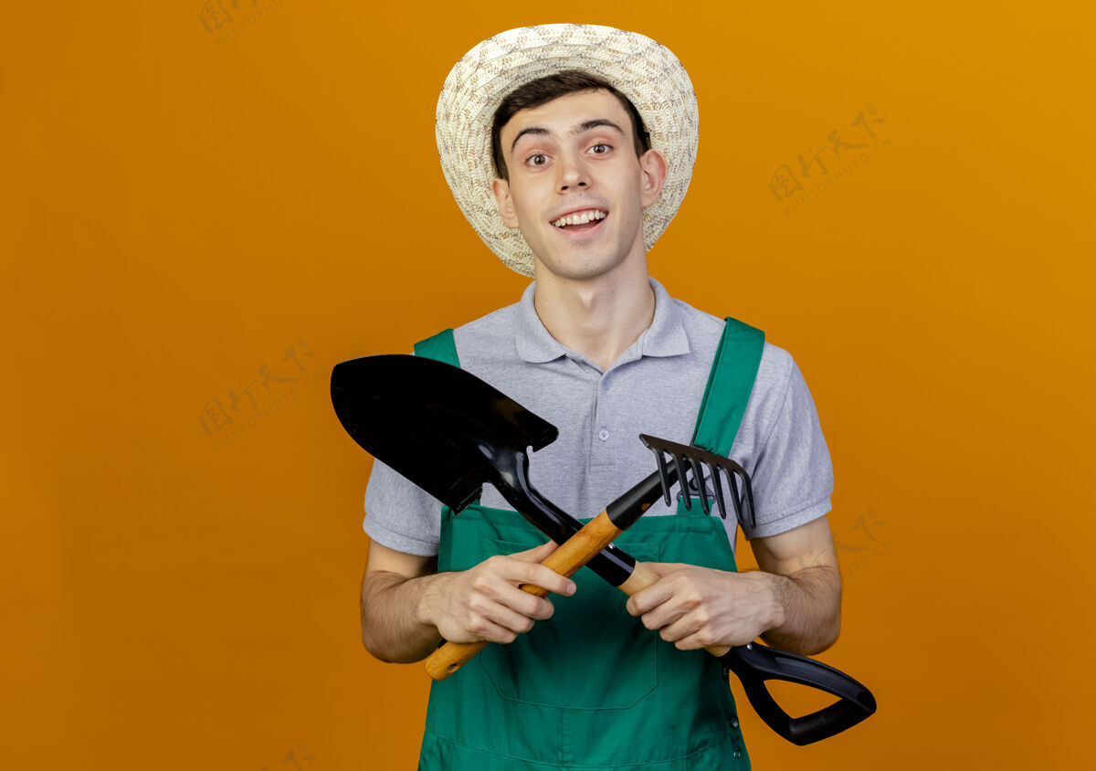 微笑微笑的年轻男园丁戴着园艺帽越过铲子和耙子耙花园帽子