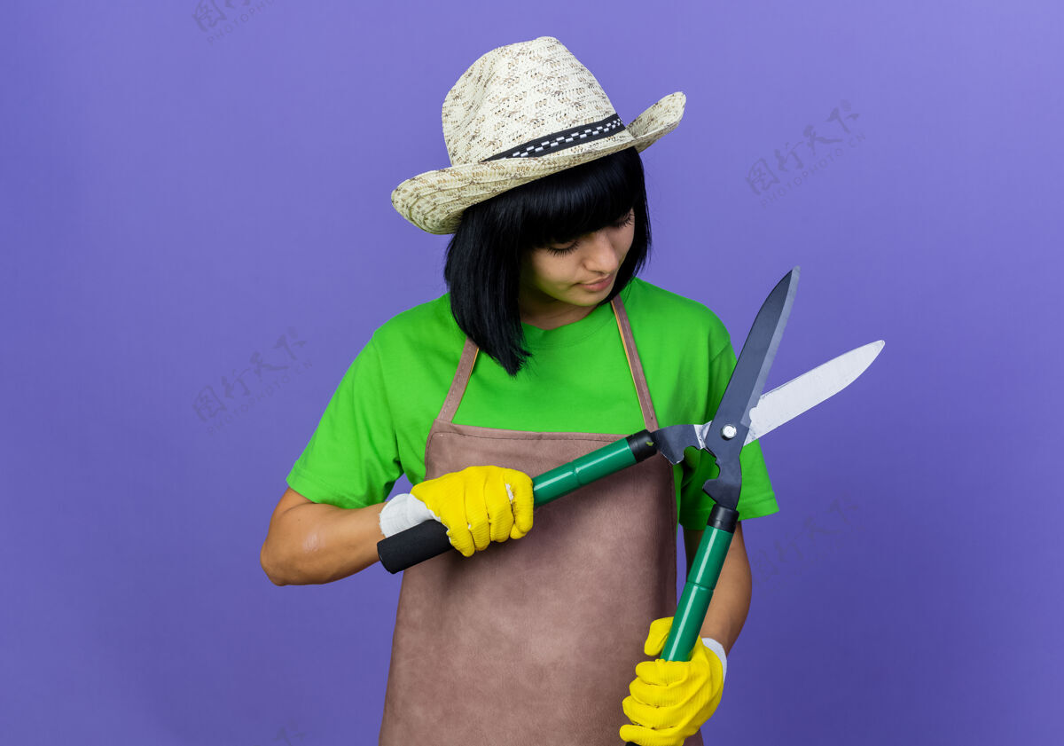 年轻穿着制服 戴着园艺帽和手套的年轻女园丁高兴地拿着剪刀看紫色复制手套