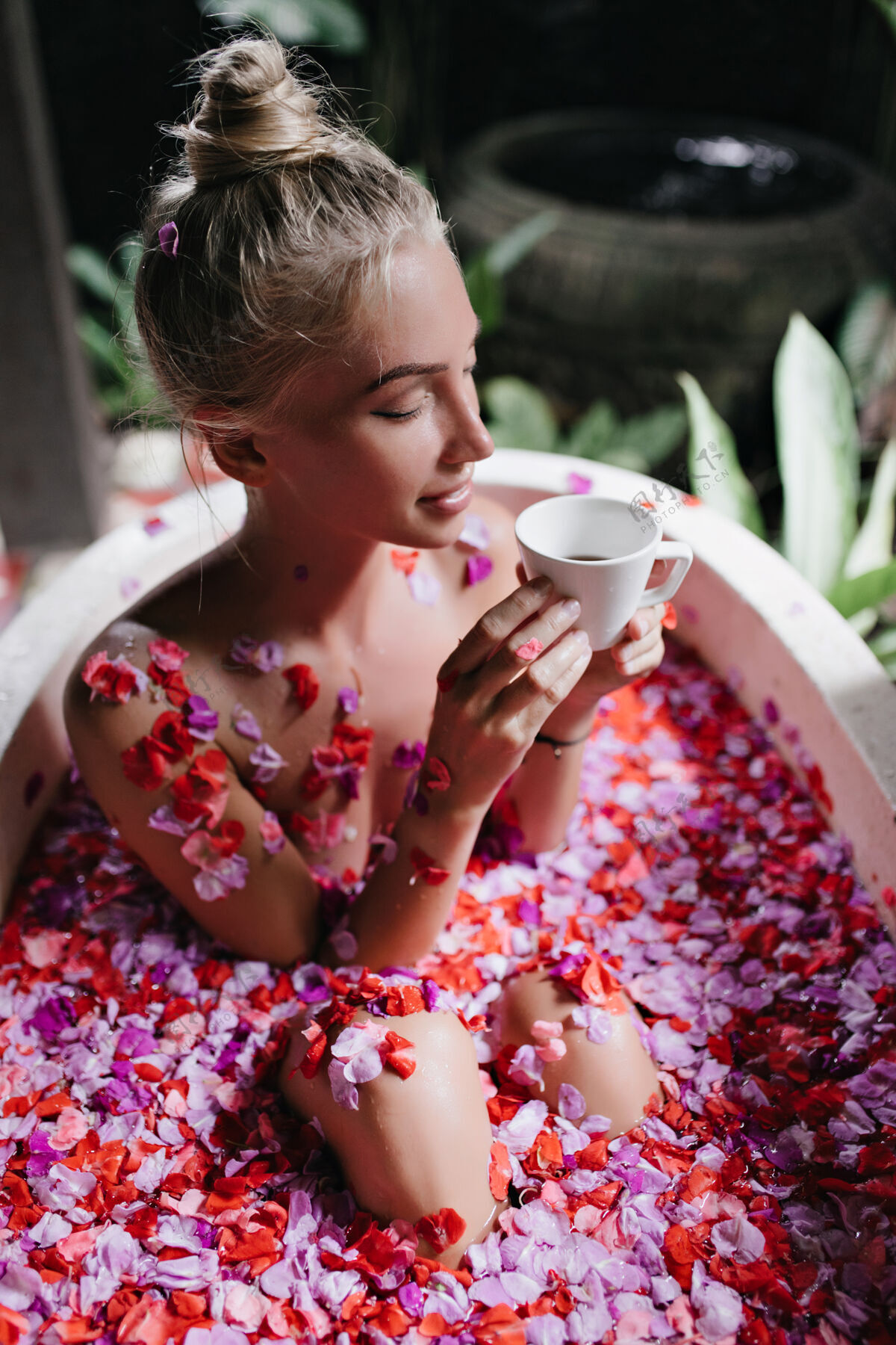 护理积极的金发女人坐在浴池里拿着鲜花和杯子浪漫的棕褐色女人在水疗时喝茶的肖像年轻沐浴快乐
