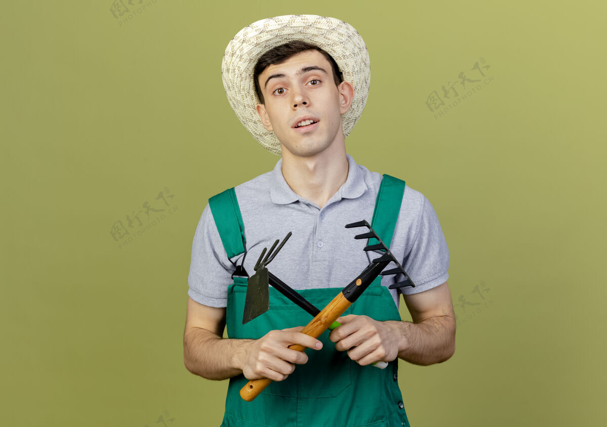 花园自信的年轻男园丁戴着园艺帽 穿过耙子和锄头耙子男性帽子信心