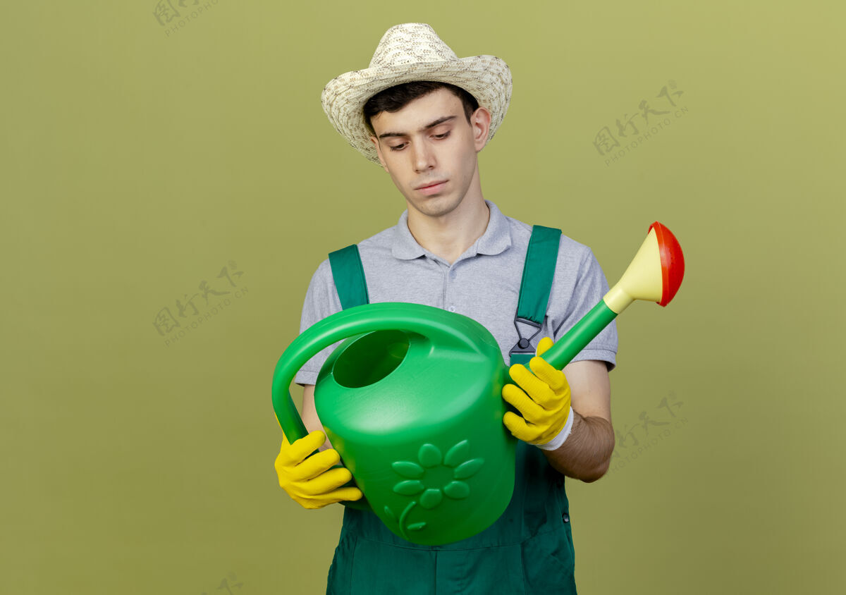橄榄自信的年轻男园丁戴着园艺帽和手套 拿着并看着浇水罐复制帽子浇水