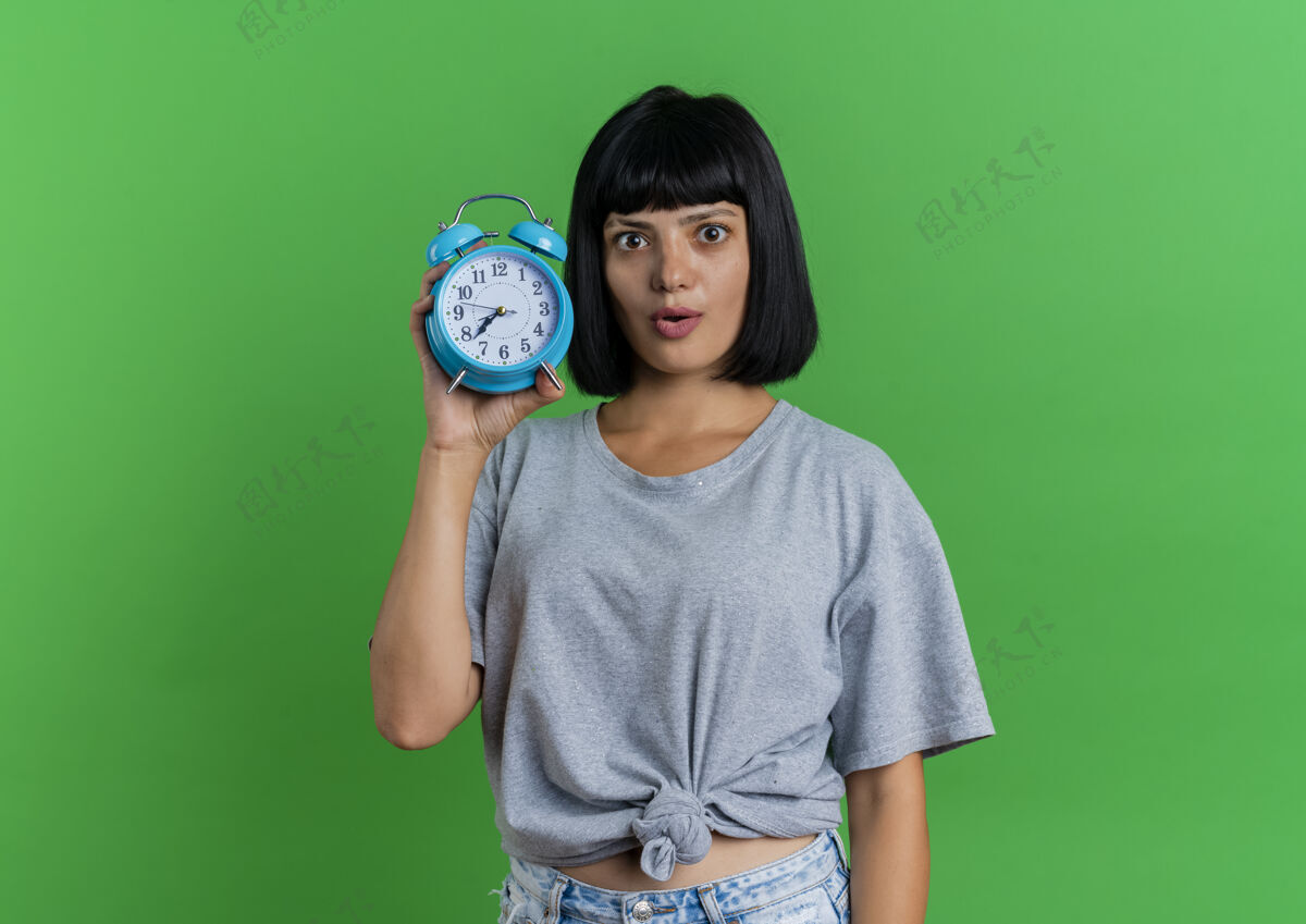 时钟兴奋的年轻黑发白种女人拿着闹钟在绿色背景上与复制空间隔离背景黑发持有