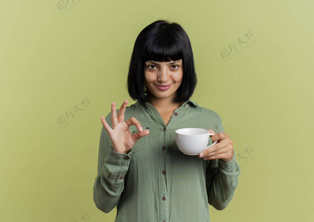 黑发高兴的年轻黑发白种女人拿着杯子 手势ok手势孤立在橄榄绿的背景和复制空间好背景年轻