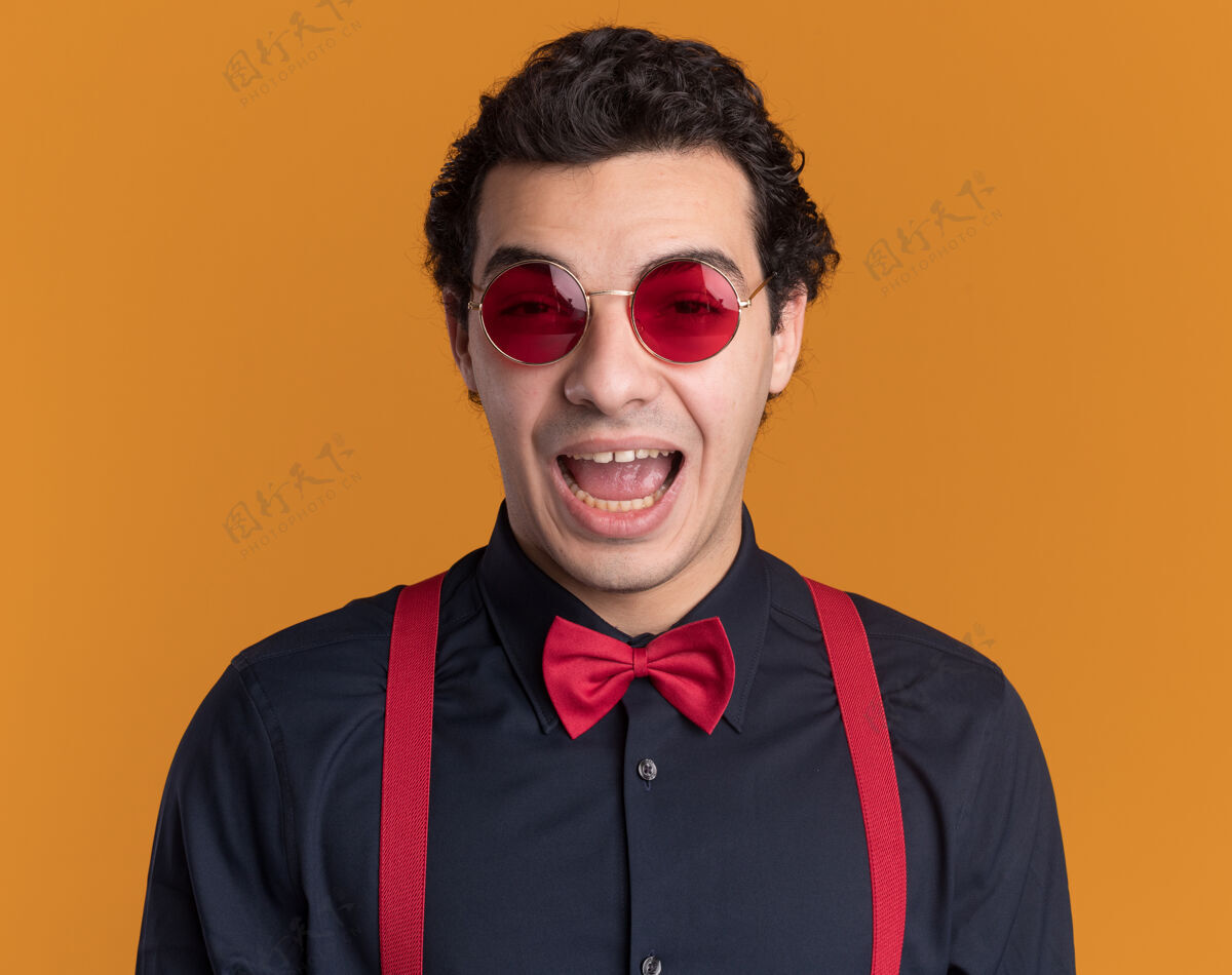 领带戴着眼镜 系着吊带 打着领结的时髦男人站在橙色的墙上 兴奋地看着前面站立穿眼镜
